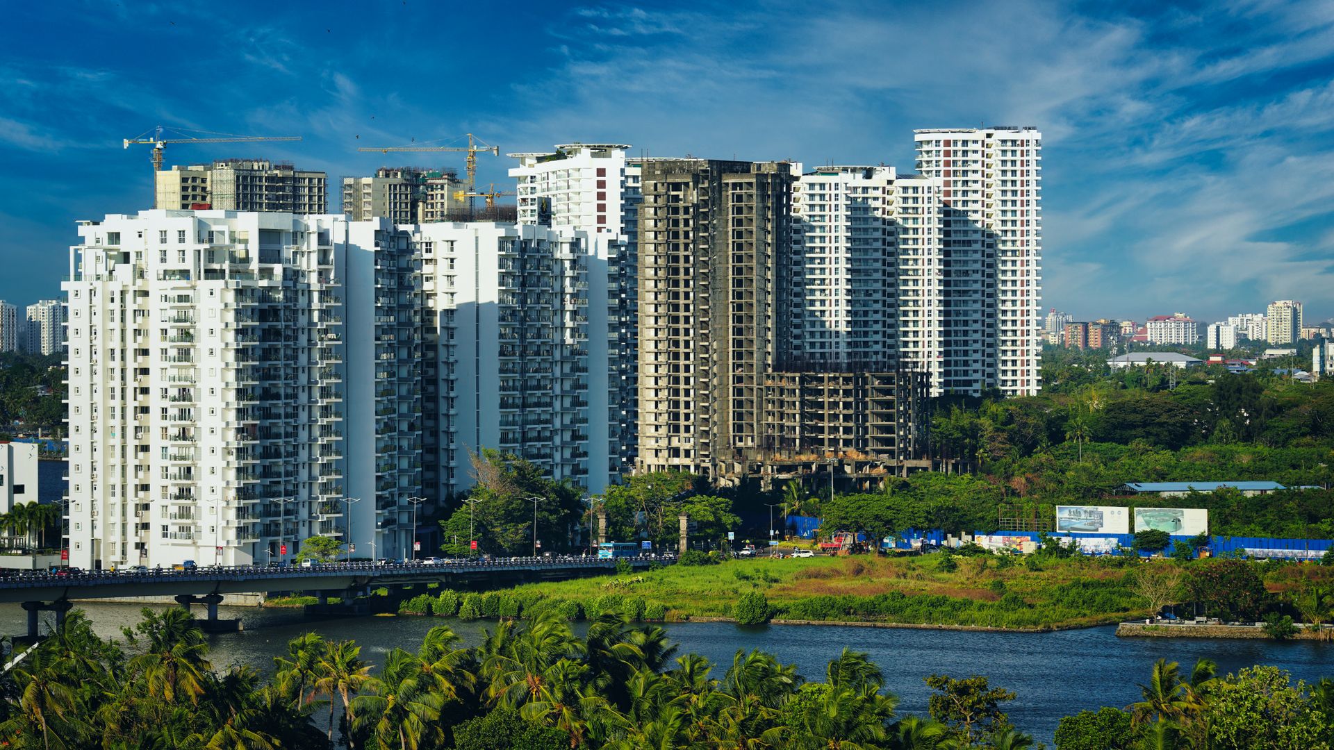 High-rise apartment towers, some under construction with cranes, above a river and palm trees in Kochi
